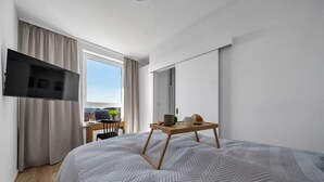 A modern hotel room with a large bed, a flat-screen TV mounted on the wall, a small desk with a cup and a plant, and a window with curtains. - 7th Floor Warsaw Włochy Apt - Balcony, Workspace - by Rentujemy (Warszawa)