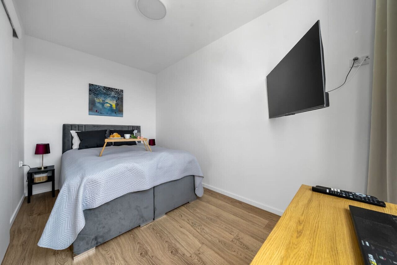 A bedroom with a bed, a bedside table, a TV mounted on the wall, and a wooden desk.