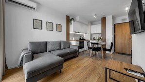 A modern living room with a grey sofa, wooden flooring, a dining area with a table and chairs, a kitchen in the background, and a mounted TV. - 7th Floor Warsaw Włochy Apt - Balcony, Workspace - by Rentujemy (Warszawa)