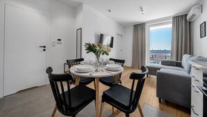 A modern living room with a dining area, a grey sofa, a TV mounted on the wall, and a large window with curtains. - 7th Floor Warsaw Włochy Apt - Balcony, Workspace - by Rentujemy (Warszawa)