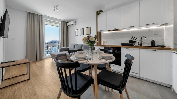 A modern kitchen with white cabinets, a marble countertop, and a dining area with a round table and black chairs. - 7th Floor Warsaw Włochy Apt - Balcony, Workspace - by Rentujemy (Warszawa)
