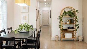 A dining area with a table, chairs, a hanging light fixture, and a shelving unit with plants.