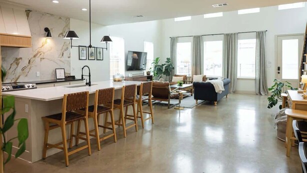 A modern kitchen with a marble backsplash, a dining area with wooden chairs, and a living room with a sofa and a TV.