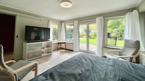 A bedroom with a bed, a TV, a desk, a chair, and a view of the garden through the window.