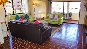 Un salon avec un canapé noir, un canapé vert et une table basse. On y trouve deux lampes, une plante en pot et une fenêtre donnant sur des immeubles.