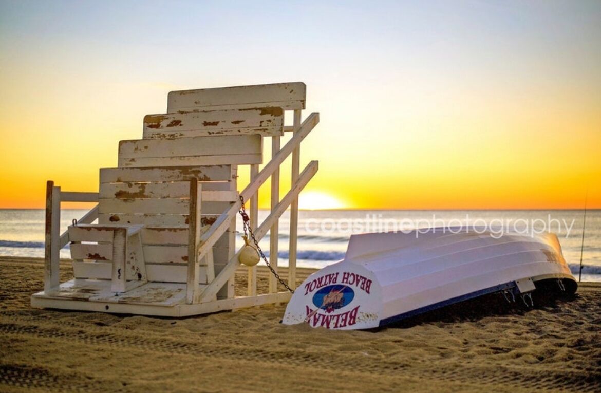 Une chaise de plage et un poste de surveillance des sauveteurs, avec un panneau, le tout sur fond de coucher de soleil.