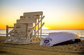 A beach chair and a lifeguard stand with a sign, set against a sunset.
