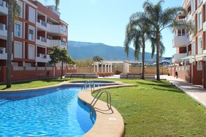 A swimming pool with a curved edge, surrounded by a well-maintained lawn and palm trees. - Apartamento CON Gran Terraza Y Acceso Directo A Piscina (Dénia)