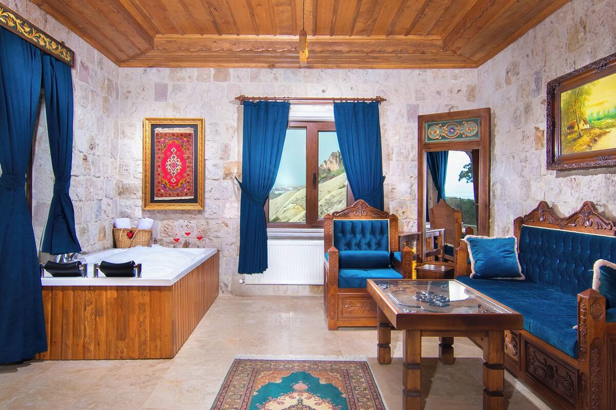 A room with a stone wall, wooden ceiling, a large window with curtains, a sofa, a wooden table, and a small bed.