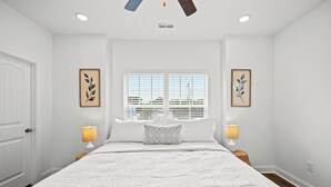 Ein Schlafzimmer mit einem großen Bett, zwei Nachttischlampen, einem Fenster mit Jalousien und zwei gerahmten Blätterbildern an den Wänden.