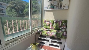 A balcony with a white table, a blue bowl, and a green pitcher. There is a couch with a leafy pattern and a wall-mounted shelf with plants. - Mise au Vert F2 à 5 mns des Commerces et 2 mns des Thermes Mer et la Montage (Amélie-les-Bains-Palalda)