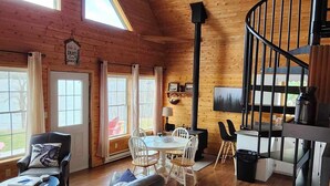 Un salón espacioso con techo alto, paredes de madera, una escalera espiral negra, chimenea y un comedor con mesa y sillas.