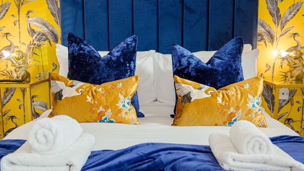 Una cama bien hecha con almohadas florales azules y amarillas, sábanas blancas y dos toallas enrolladas sobre un edredón azul.