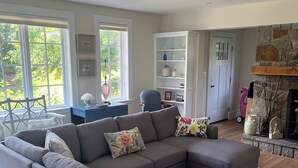 A living room with a sectional sofa, a dining area with a table and chairs, a fireplace, and a view of greenery outside. - Plum Cove Beach Paradise. Overlook Plum Cove from your kitchen n only steps away (Gloucester)