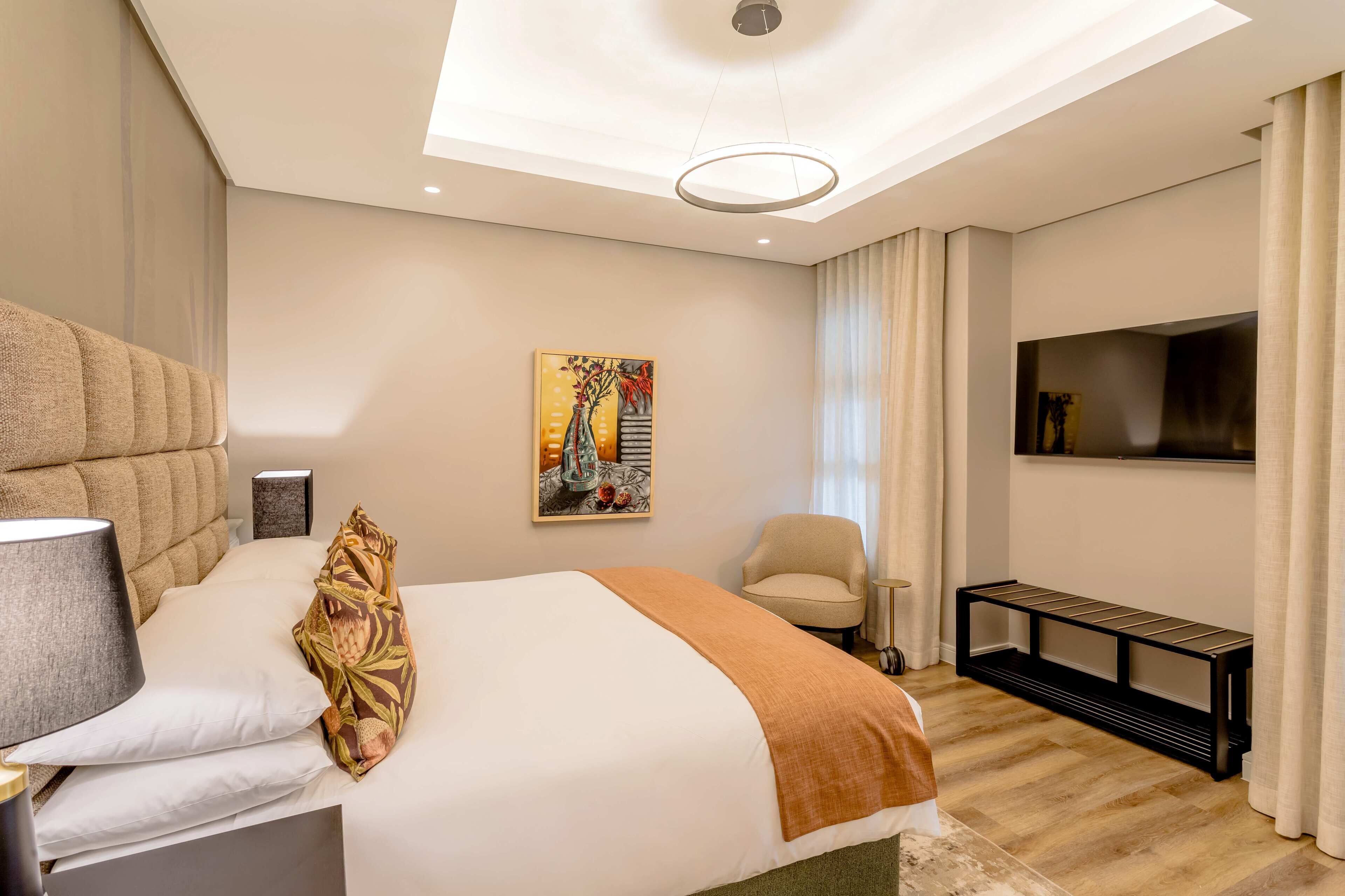 A hotel room with a bed, a chair, a TV, a painting, and a bench.