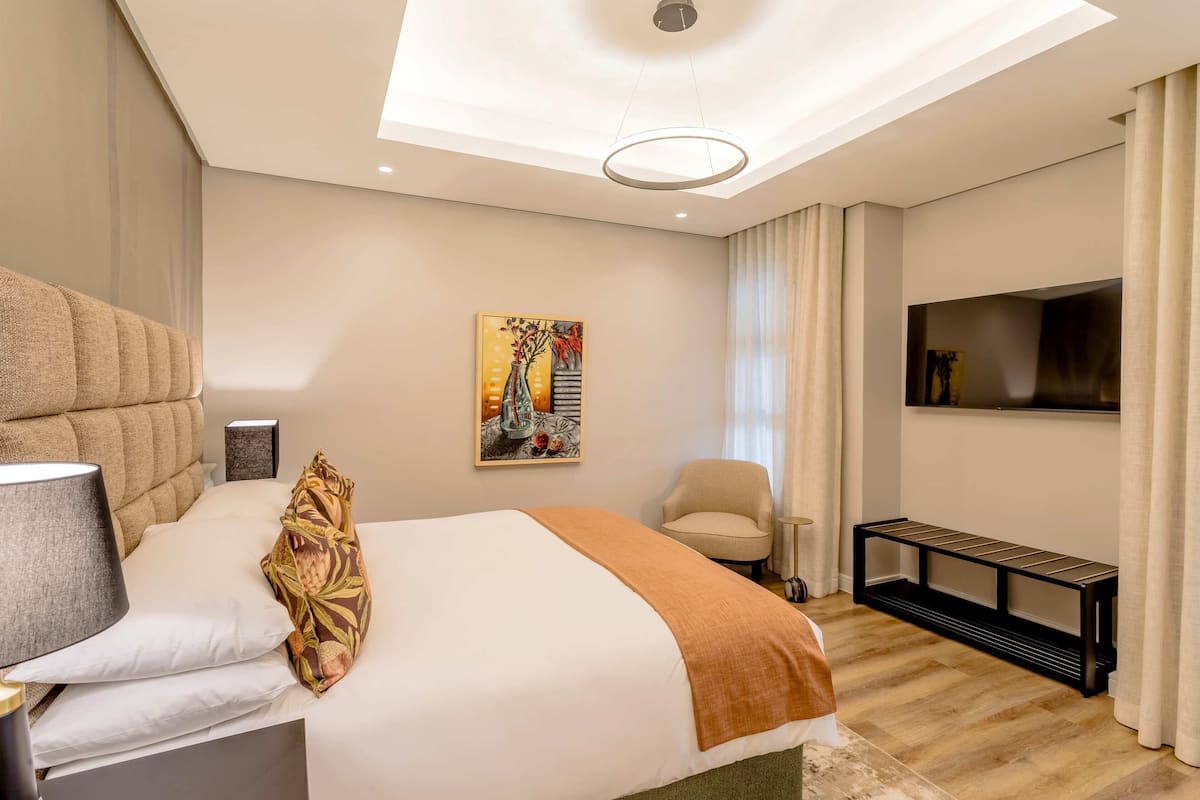 A hotel room with a bed, a chair, a TV, a painting, and a bench.