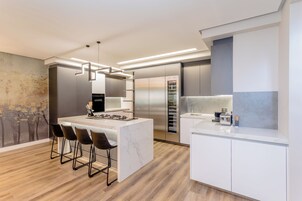 A modern kitchen with a central island, bar stools, stainless steel appliances, and a large window.