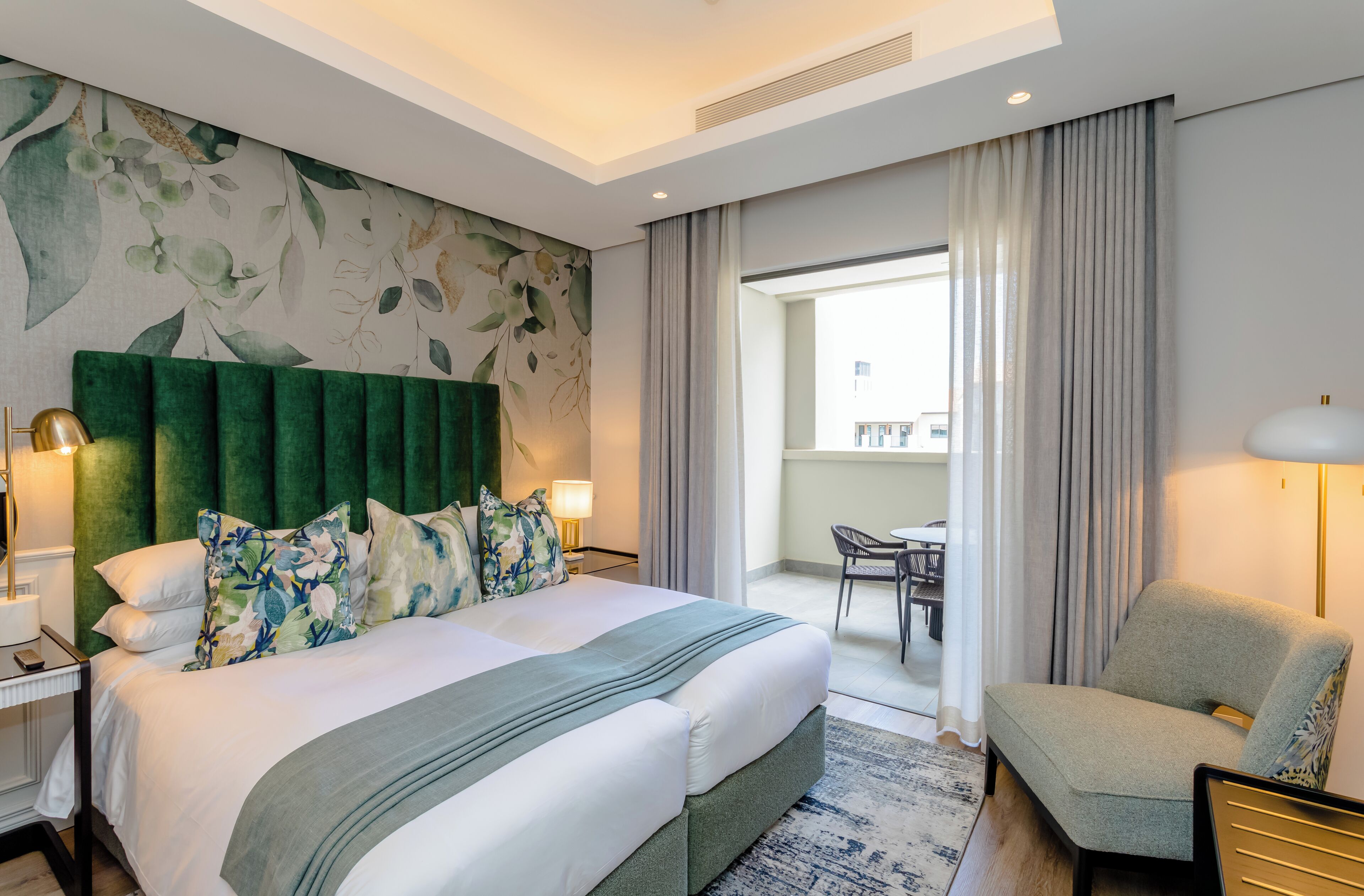 Habitación de hotel con una cama grande, un cabecero verde, almohadas estampadas con motivos florales, una mesita de noche con una lámpara y vistas al balcón.