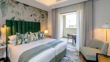 Habitación de hotel con una cama grande, un cabecero verde, almohadas estampadas con motivos florales, una mesita de noche con una lámpara y vistas al balcón.
