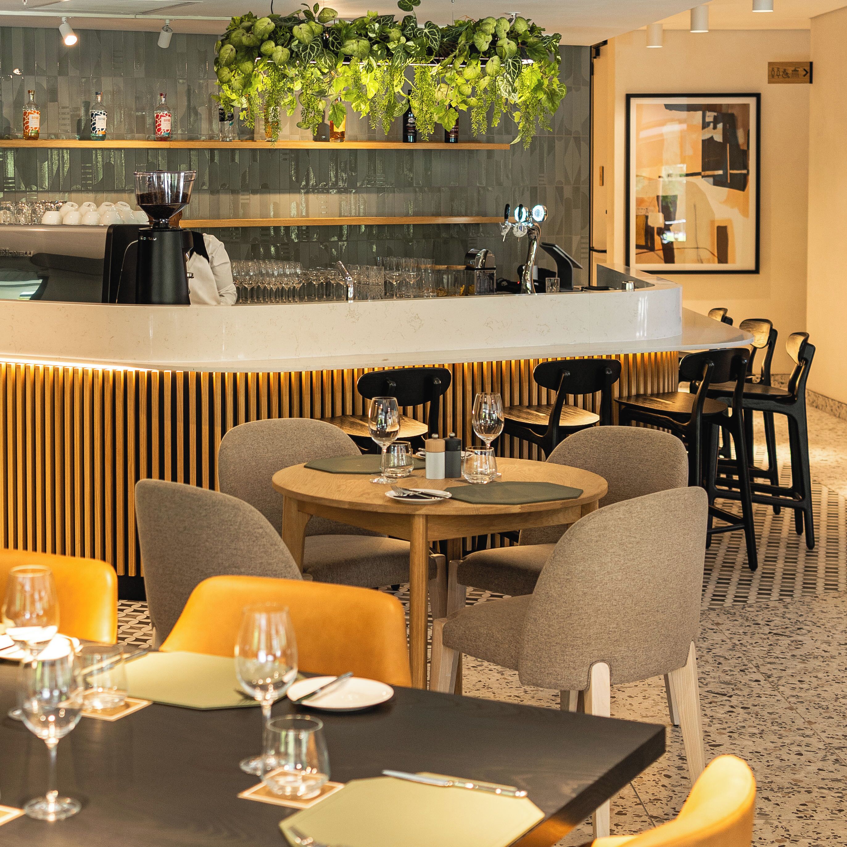 Un restaurante con mesas redondas, sillas y una zona de bar con mostrador y plantas colgantes.