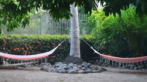 Un hamac est suspendu entre deux arbres dans un jardin, avec une bordure de pierres et une clôture en arrière-plan.
