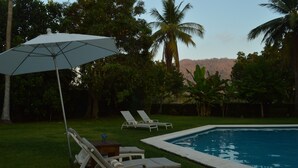 Un espace aménagé au bord de la piscine, avec deux chaises longues et un parasol, entouré d’une végétation luxuriante et de palmiers.