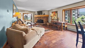 Un salon avec une cheminée, un canapé, une table à manger et une grande fenêtre donnant sur des immeubles et des montagnes.