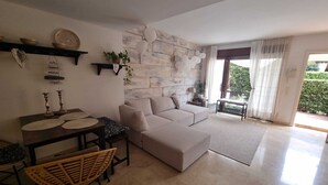 A modern living room with a stone accent wall, a sofa, a dining table, and a view of greenery outside. - Casa Violette (Benissa)
