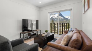 A modern living room with a flat-screen TV, a dark wooden coffee table, a leather sofa, and a large window offering a mountain view.