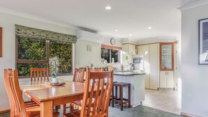 A spacious dining area with a wooden table and chairs, a kitchen in the background, and a view of greenery outside. - Moetapu Magic - Marlborough Sounds Holiday Home (Picton)