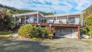 A modern two-story house with a covered deck, a garage, and a scenic hillside backdrop. - Moetapu Magic - Marlborough Sounds Holiday Home (Picton)