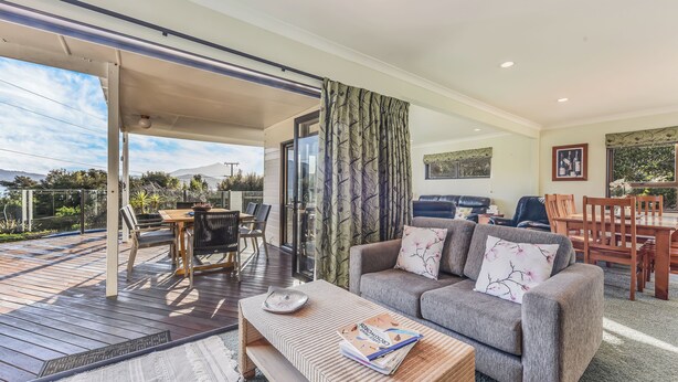 A spacious living area with a grey sofa, wooden coffee table, and a dining table with chairs. A large window offers a view of the outdoors. - Moetapu Magic - Marlborough Sounds Holiday Home (Picton)
