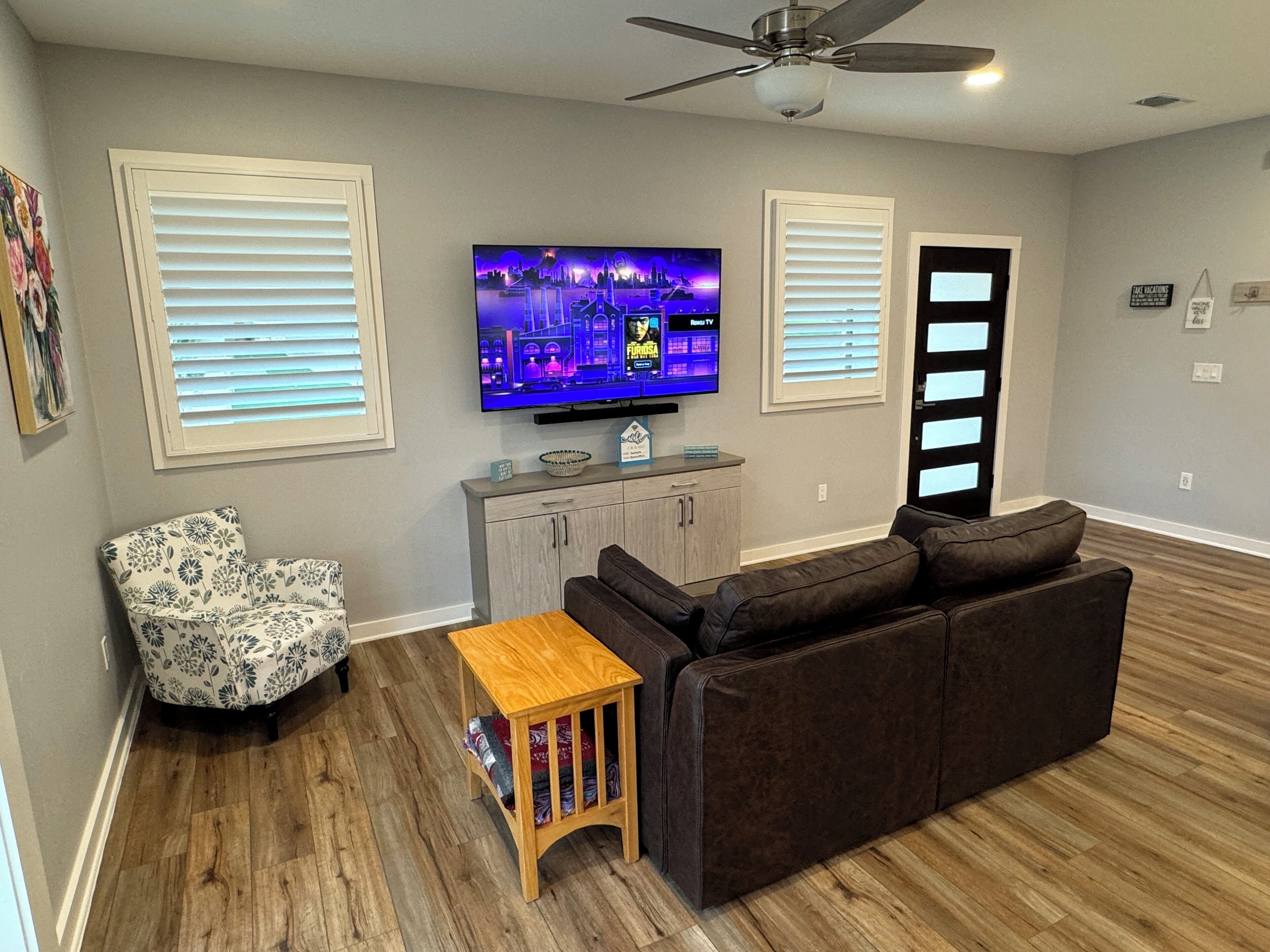 A living room with a flat-screen TV, a brown sofa, a patterned armchair, a small wooden table, and a ceiling fan.