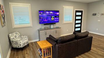 A living room with a flat-screen TV, a brown sofa, a patterned armchair, a small wooden table, and a ceiling fan.