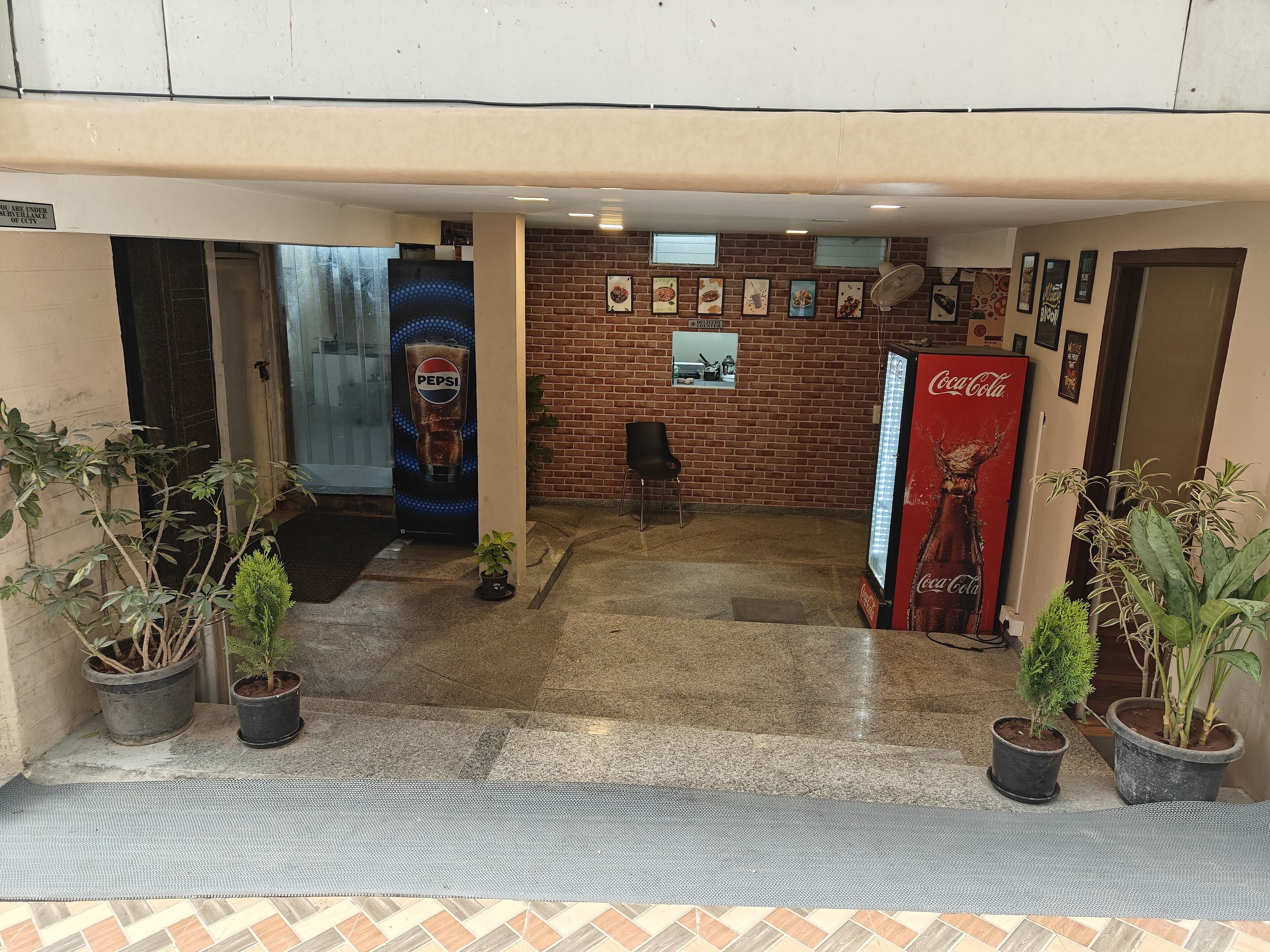 A lobby with a brick wall, a Pepsi advertisement, a Coca-Cola refrigerator, and potted plants.