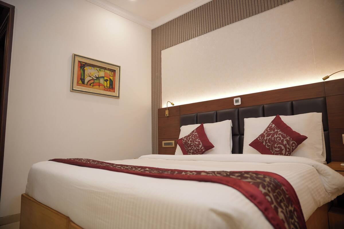 A hotel room with a neatly made bed, a headboard with built-in lighting, a nightstand, and a wall-mounted artwork.