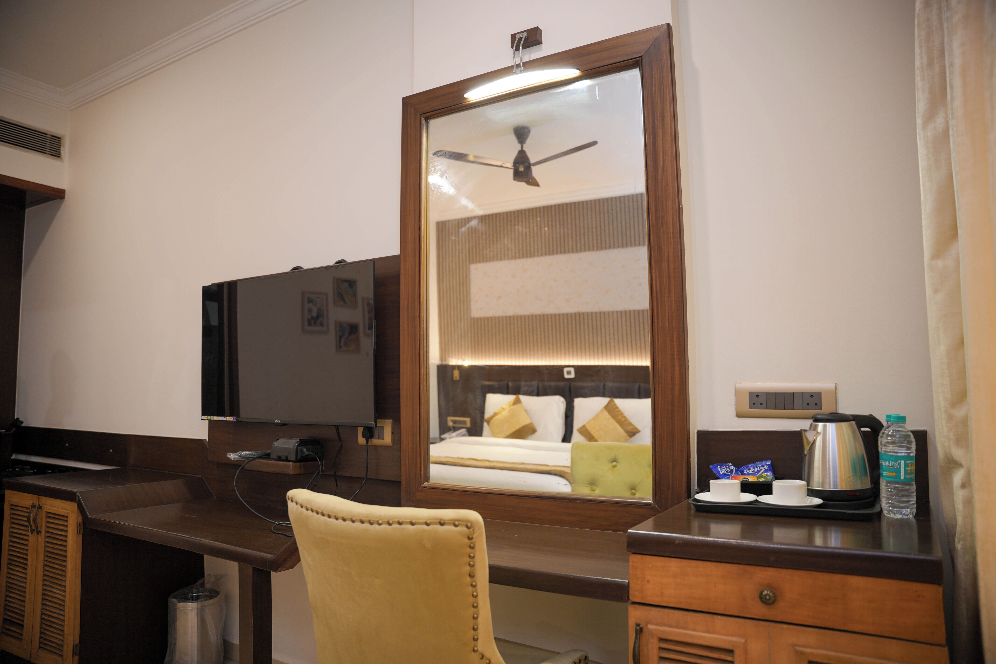 A hotel room with a wooden desk, a television, a mirror, a bed with pillows, a bedside table with a kettle, cups, and a bottle of water.