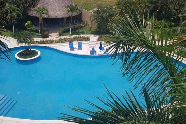 A large swimming pool with lounge chairs and umbrellas, surrounded by palm trees and a thatched-roof building.