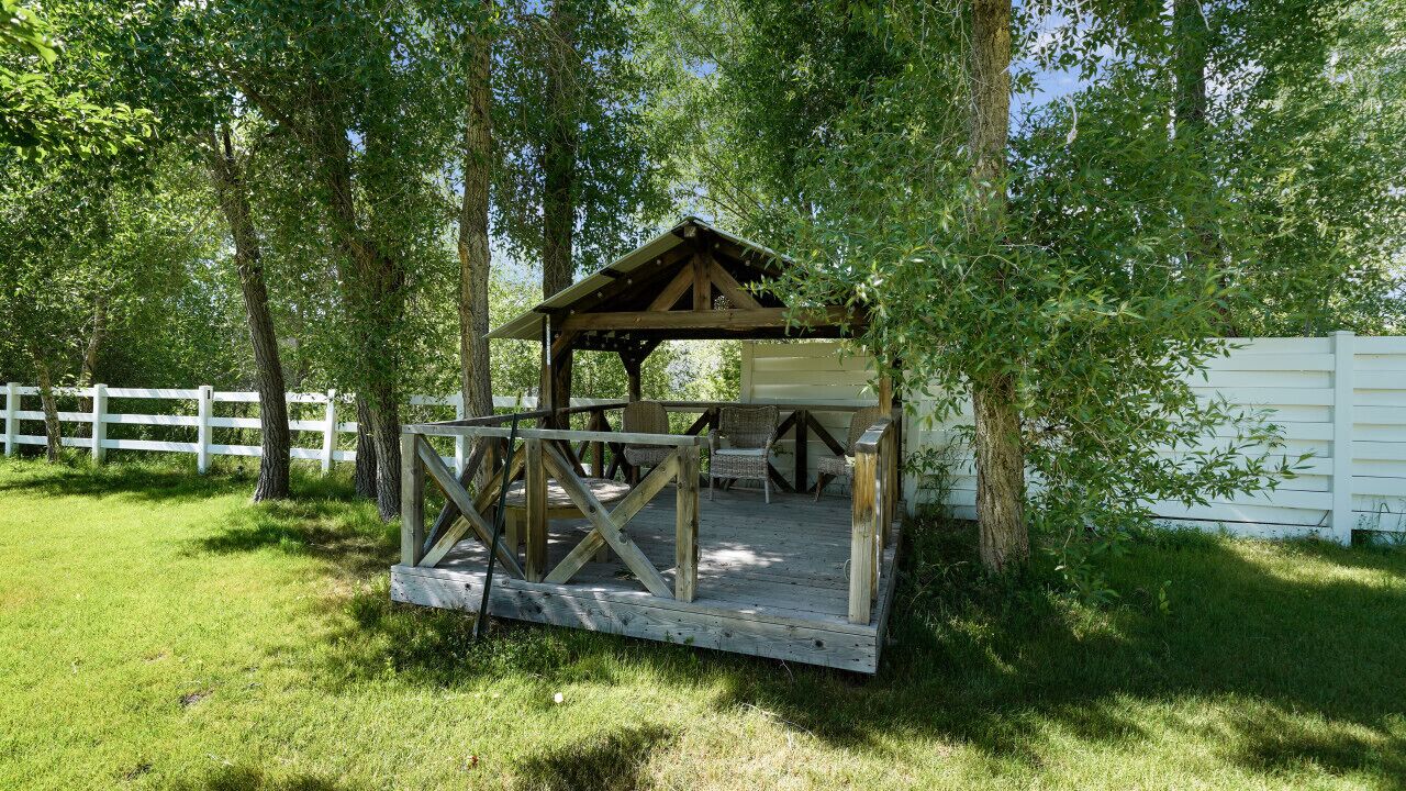 A wooden gazebo with a bench and table, surrounded by trees and a white fence.
