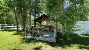 A wooden gazebo with a bench and table, surrounded by trees and a white fence.