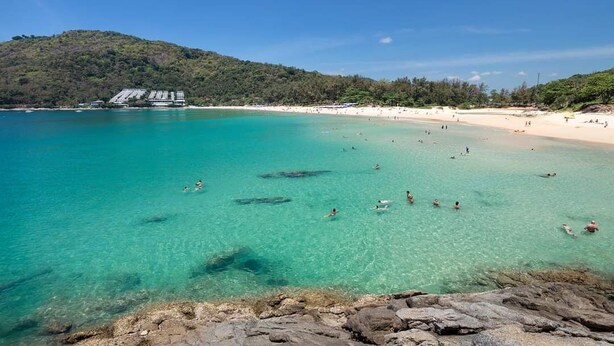 Ein Strand mit klarem türkisblauem Wasser, einem felsigen Vordergrund und einem Gebäude mit geneigtem Dach im Hintergrund.