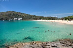 En strand med kristallklart turkost vatten, en stenig förgrund och en byggnad med ett sluttande tak i bakgrunden.
