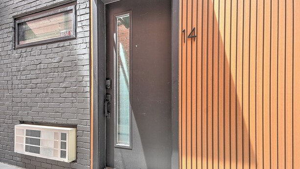 Ein moderner Gebäudeeingang mit Glastür, kleinem Fenster und Lüftungsgitter an der Backsteinwand.