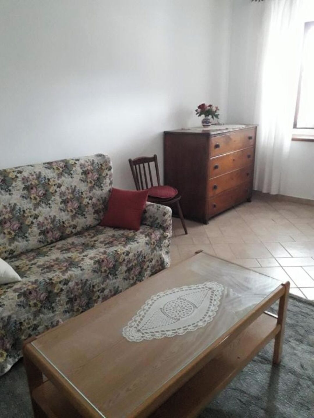 Un salon avec un canapé à motifs floraux, une table basse en bois surmontée d’un chemin de table en dentelle, une chaise en bois, une commode en bois et une fenêtre avec des rideaux.