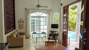 Ampla sala de estar com sofá, mesa de jantar e cadeiras. Uma televisão está fixada na parede, e há uma grande janela com vista para uma piscina.