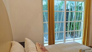 Quarto com uma janela grande, cama com almofadas brancas e listradas, e vista para palmeiras lá fora.