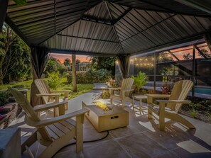 Een terras buiten met Adirondack-stoelen, een vuurplaats en een overdekt gedeelte met uitzicht op een zwembad en de ondergaande zon.