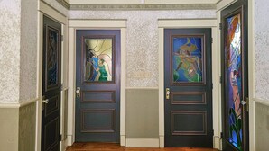 Um corredor com duas portas de madeira escura, cada uma com um vitral. As paredes têm textura e há duas luminárias no teto.