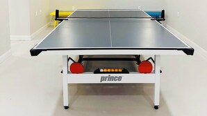 Una mesa de ping pong con estructura blanca y detalles rojos, ubicada en una habitación con paredes blancas e iluminación empotrada.