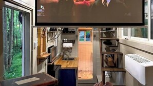 A cozy room with a large flat-screen TV mounted on the ceiling, a wooden chest labeled "Harry Potter," and a window with a view of a forest.